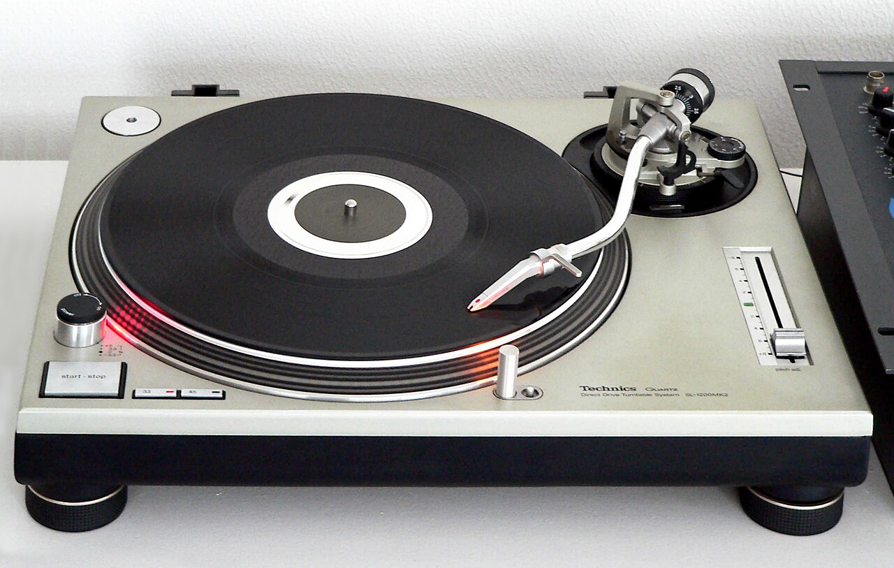 Technics SL-1200MK2 casi cenital, con plato, brazo, cápsula, start/stop, velocidades, pitch y luz de aguja visibles.